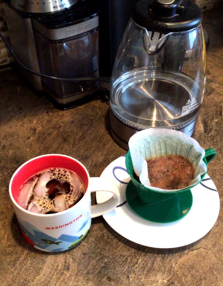 mug of Pour over coffee with ice on a brown counter with a used cone filter on a plate next to it