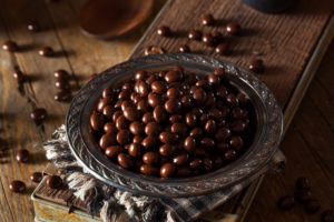 A bowl of chocolate covered nuts alongside a cold brew coffee on a wooden table.