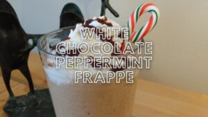 white chocolate peppermint frappe in a clear glass garnished with whipped cream, chocolate syrup and a candy cane on a wooden table.