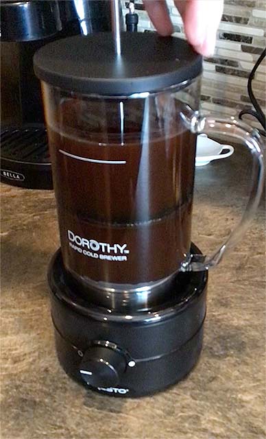pressing the plunger down on the dorothy presto cold brewer to filter the coffee, plunger is halfway down the carafe and the carafe is on the base. Brown countertop.
