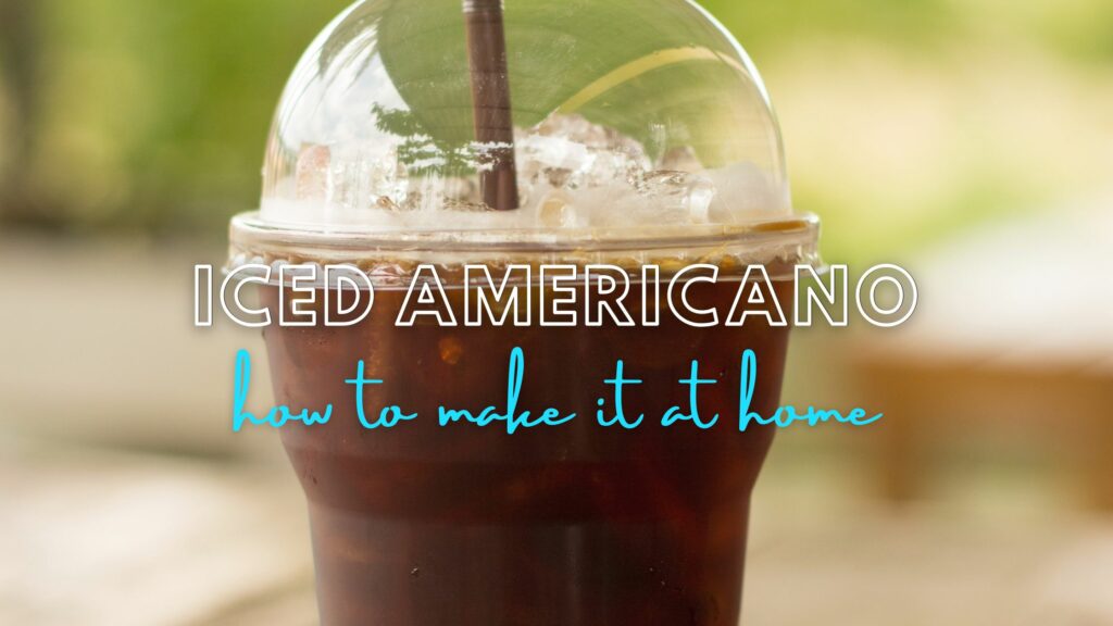 iced americano drink in a clear plastic cup with a domed lid and brown straw. On a blurry table. Text is Iced Americano How to Make it At Home.